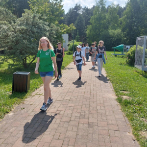 Spacer po terenie Zoom Natury – uczestnicy wycieczki edukacyjnej przemieszczają się alejką otoczoną zielenią. Na pierwszym planie widoczna jest przewodniczka w zielonej koszulce, za nią uczniowie, którzy uśmiechnięci i zaciekawieni kontynuują zwiedzanie interaktywnego parku nauki. Po prawej stronie kadru znajduje się fragment placu zabaw. Atmosfera zdjęcia oddaje letnią pogodę i entuzjazm grupy.